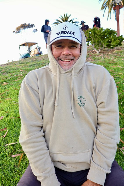 Man wearing a beige hoodie and cap with 'Yardage' branding, sitting on grass.