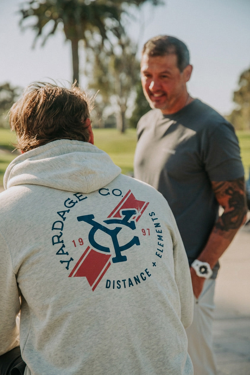 A man wearing a light gray hoodie with a large navy and red 'Ardage Co' graphic on the back, standing outdoors and talking to another man in a dark gray t-shirt.