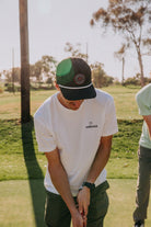 A man on a golf course wearing a white YARDAGE t-shirt, dark green pants, and a black and green trucker hat, looking down while playing.