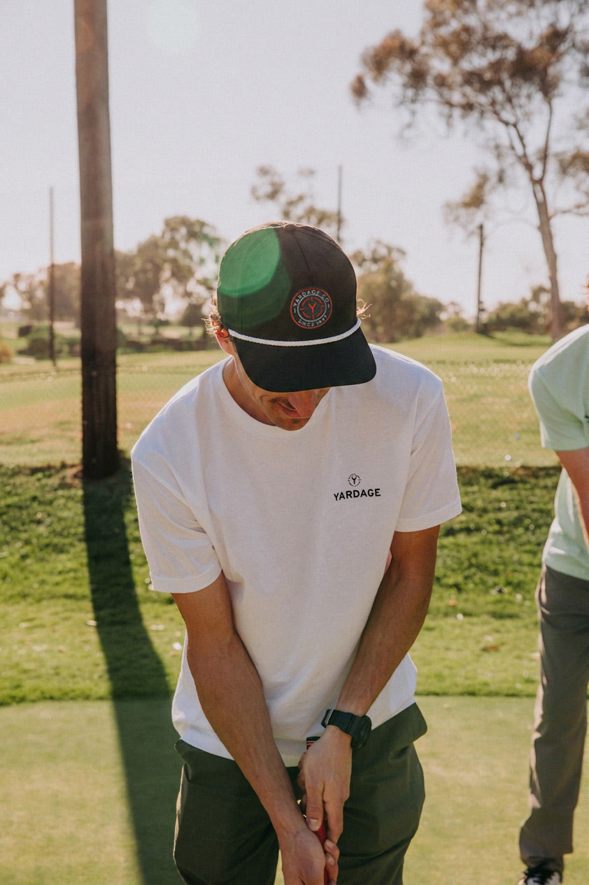 A man on a golf course wearing a white YARDAGE t-shirt, dark green pants, and a black and green trucker hat, looking down while playing.