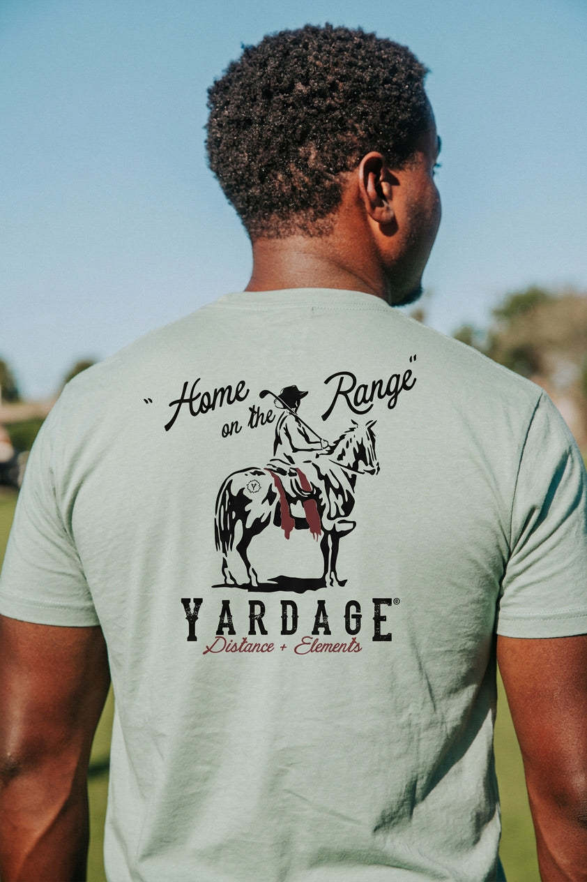 A man viewed from behind wearing a sage green t-shirt with a black graphic print of a cowboy riding a horse and the text 'Home on the Range' and 'YARDAGE Distance Elements'.