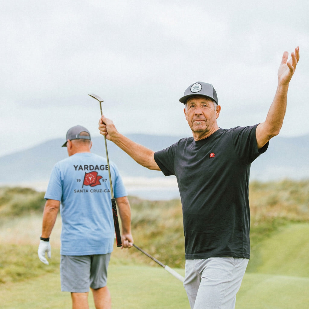 A man on a golf course raises his arm while holding a golf club, wearing a black t-shirt and light gray pants. Another man in a light blue t-shirt walks away in the background under an overcast sky.