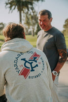 A man wearing a light gray hoodie with a large navy and red 'Ardage Co' graphic on the back, standing outdoors and talking to another man in a dark gray t-shirt.